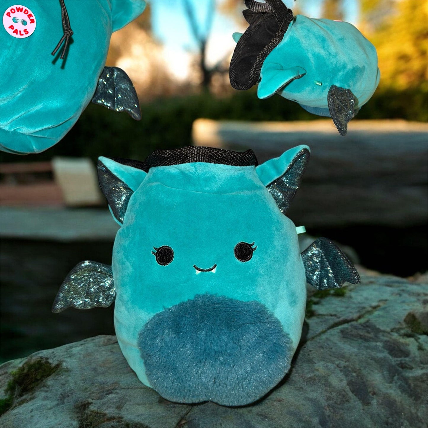 Teal coverted plush toy into handmade chalk bag with black glittery wings on an nature background flying bat bags with "Powder Pals" button logo